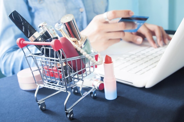 Sección media de una mujer usando una computadora portátil con productos de belleza en el carrito de compras en la mesa