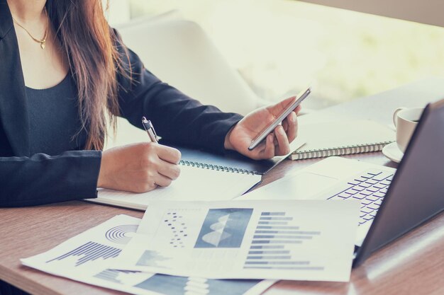 Foto sección media de una mujer usando el teléfono mientras escribe en un libro con una computadora portátil en la mesa