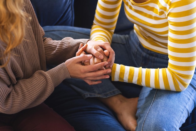 Sección media de una pareja de lesbianas biraciales tomadas de la mano sentadas en el sofá en casa