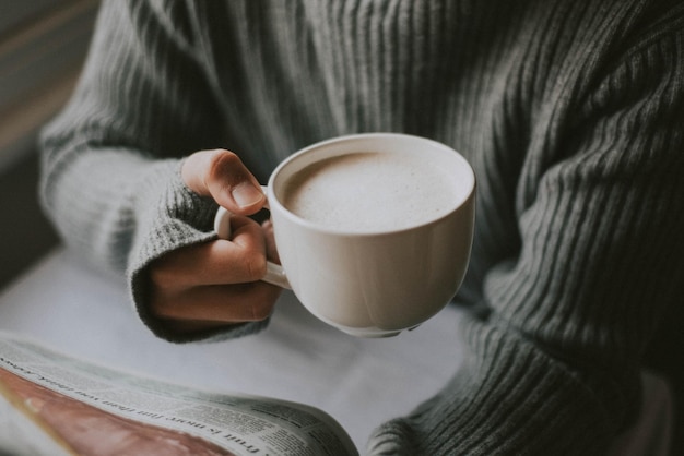 Sección media de una persona leyendo un periódico mientras sostiene una taza de café en la mesa