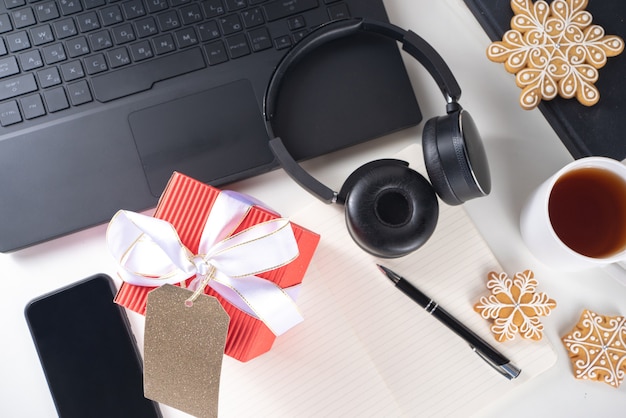 Secret Santa. Celebração de Natal no escritório. Mesa de escritório com laptop, blocos de notas, material de escritório, smartphone e uma grande caixa de presente de Natal com laço festivo. flatlay