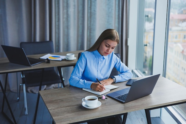 La secretaria revisa el horario Alegre joven hermosa mujer con gafas con una sonrisa usa una computadora portátil mientras está sentada en su lugar de trabajo