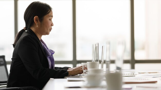 Secretário asiático, de meia idade, feliz confiante feminino empresária bem-sucedida em terno formal, sentado na mesa de trabalho, digitando informações no computador laptop laptop sozinho na sala de escritório da empresa em silêncio.