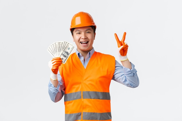 Sector de la construcción y concepto de trabajadores industriales. Feliz sonriente constructor asiático, gerente de construcción con casco y ropa reflectante que muestra el signo de la paz y dinero, salario, pared blanca.