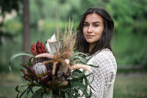 Seductora joven morena con un vestido blanco con un ramo de flores en el bosque sobre un fondo borroso,