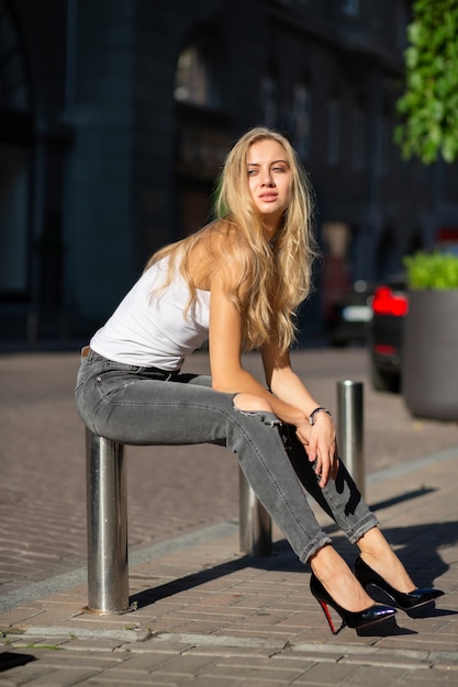 Seductora modelo rubia con cabello largo con jeans y camiseta posando en la ciudad bajo los rayos del sol