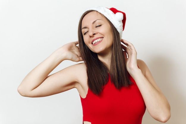 Seductora mujer caucásica joven feliz con los ojos cerrados con placer y sonrisa encantadora con vestido rojo y sombrero de Navidad sobre fondo blanco. Niña de Santa aislada de cerca. Vacaciones de año nuevo 2018