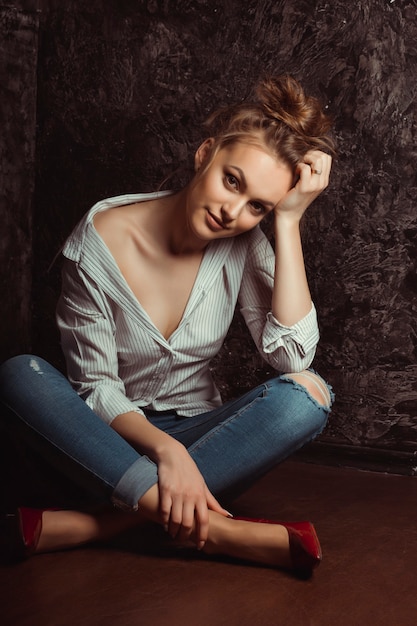 Seductora mujer joven en camisa y jeans sentados en el suelo en el estudio
