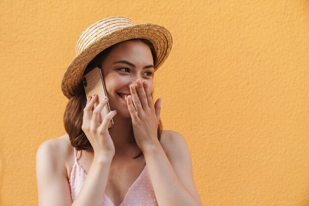 Foto seductora mujer joven con sombrero de paja de verano sonriendo y hablando por teléfono celular aislado en amarillo