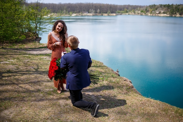 See im Wald: Ein Mann überrascht seine Freundin, er steckt ihr einen Ring an den Finger, eine Liebeserklärung und ein Heiratsangebot. Mädchen mit einem Lächeln im Gesicht und einem Strauß roter Rosen