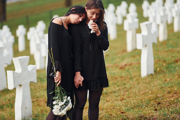 Segura flores Duas jovens em roupas pretas visitando cemitério com muitas cruzes brancas Concepção de funeral e morte