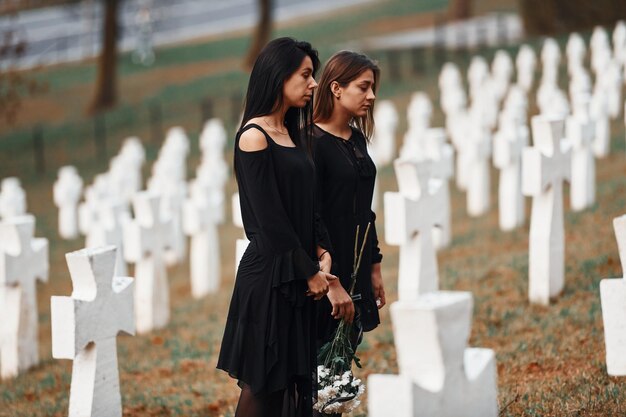 Segura flores Duas mulheres jovens em roupas pretas visitando cemitério com muitas cruzes brancas Concepção de funeral e morte