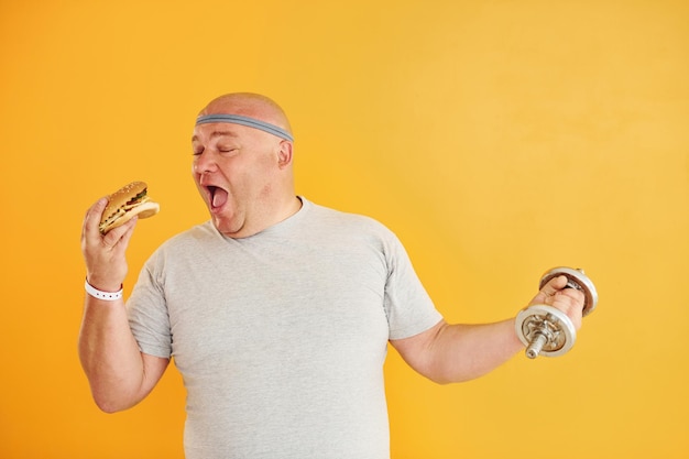 Segura hambúrguer e haltere Homem engraçado com excesso de peso em gravata esportiva contra fundo amarelo