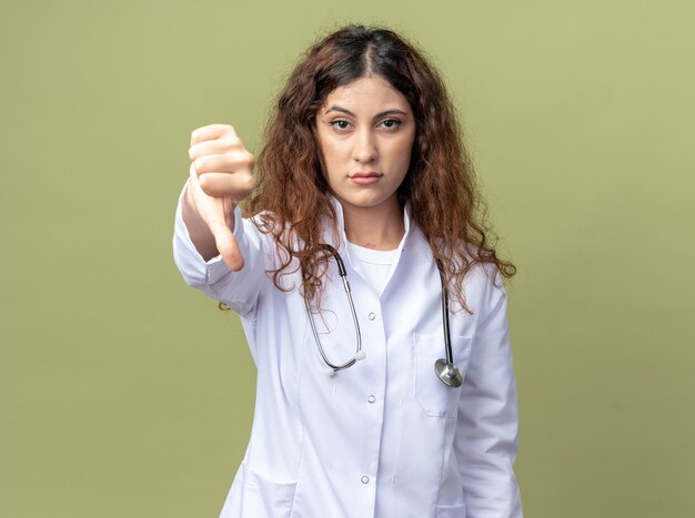 Segura joven doctora vistiendo bata médica y un estetoscopio mirando al frente mostrando el pulgar hacia abajo aislado en la pared verde oliva