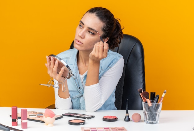 Segura de mujer bastante caucásica sentada a la mesa con herramientas de maquillaje sosteniendo espejo y aplicando sombra de ojos con pincel de maquillaje mirando al lado