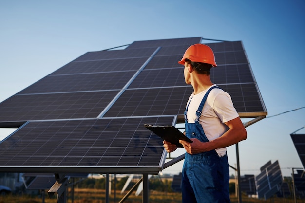 Segura o bloco de notas na mão. Trabalhador masculino com uniforme azul ao ar livre com baterias solares em dia ensolarado.