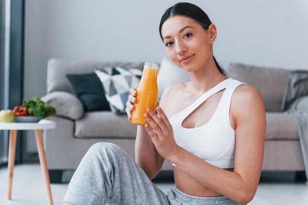 Segura o copo com suco de laranja nas mãos Jovem mulher com corpo esbelto em roupas esportivas tem dia de fitness dentro de casa