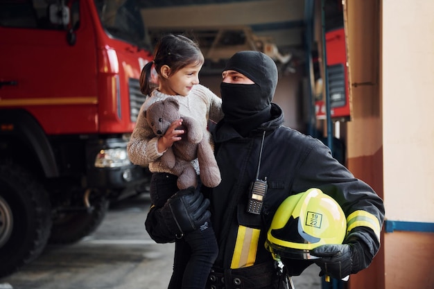 Segura o ursinho de pelúcia A garotinha feliz está com bombeiro masculino em uniforme de proteção