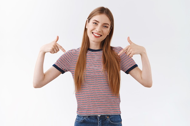 Segura y orgullosa, jactanciosa mujer guapa con camiseta a rayas sonriendo segura de sí misma, inclinando la cabeza y mirando a la cámara como promoviendo sus propios logros, señalando a sí misma fanfarroneando, fondo blanco