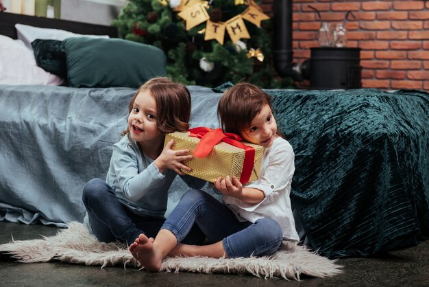 Segurando aquela caixa amarela e interessado no que está dentro. férias de natal com presentes para essas duas crianças que estão sentadas dentro de casa no belo quarto perto da cama.