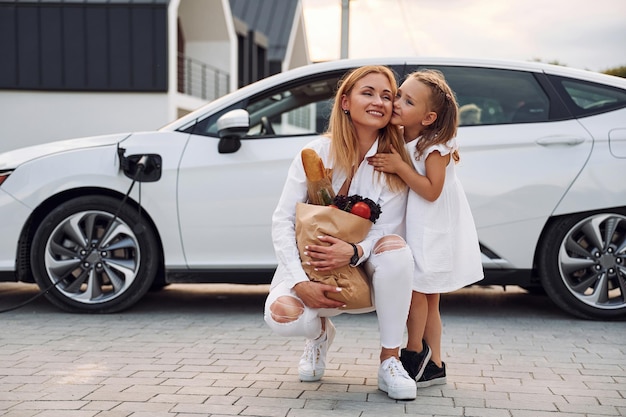 Segurando o saco com produtos Jovem mulher com sua filhinha está com seu carro elétrico ao ar livre