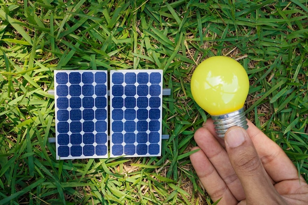 Segurando uma lâmpada ao lado de um painel solar fotovoltaico no fundo da grama Conceito alternativo de energia limpa