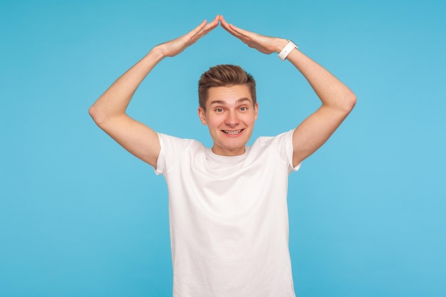 Seguridad y seguro de hogar. Retrato de un buen hombre amistoso con una camiseta blanca haciendo un gesto de techo sobre la cabeza y sonriendo a la cámara con una expresión amable. Foto de estudio interior aislado sobre fondo azul.