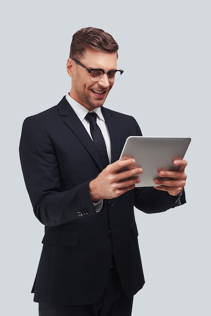 Seguro e inteligente. Hombre joven guapo con tableta digital y sonriendo mientras está de pie contra el fondo gris