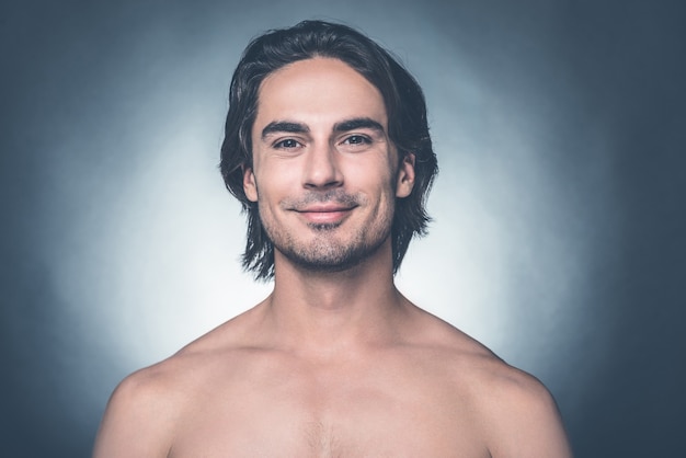 Seguro y guapo. Retrato de joven sin camisa mirando a la cámara y sonriendo mientras está de pie contra el fondo gris