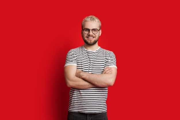 Seguro hombre caucásico con cabello rubio y barba corta posando con las manos cruzadas sobre un fondo rojo.