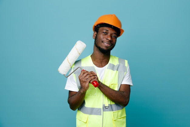 Seguro joven constructor afroamericano en uniforme con cepillo de rodillos aislado sobre fondo azul.