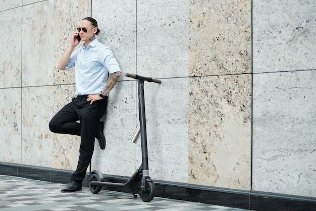 Seguro joven empresario elegante apoyado en la pared junto a su scooter y llamando por teléfono