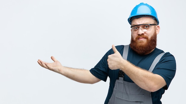 Seguro joven trabajador de la construcción masculino vistiendo uniforme de casco de seguridad y gafas de seguridad mirando a la cámara mostrando la mano vacía y el pulgar hacia arriba aislado sobre fondo blanco.