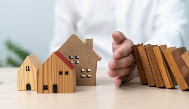 El seguro con las manos protege una casa El bloque de dominó de madera está a punto de caer sobre la casa Seguro de hogar o concepto de seguro de casa