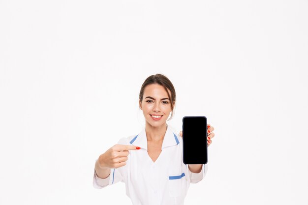 Seguro médico joven vistiendo uniforme que se encuentran aisladas sobre la pared blanca, mostrando teléfono móvil con pantalla en blanco