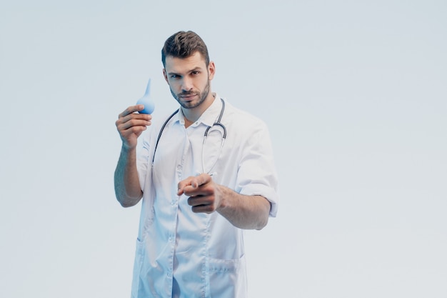 Seguro médico varón europeo muestra enema médico y apuntando con el dedo a la cámara. Joven barbudo con estetoscopio con bata blanca. Aislado sobre fondo gris. Sesión de estudio. Copia espacio