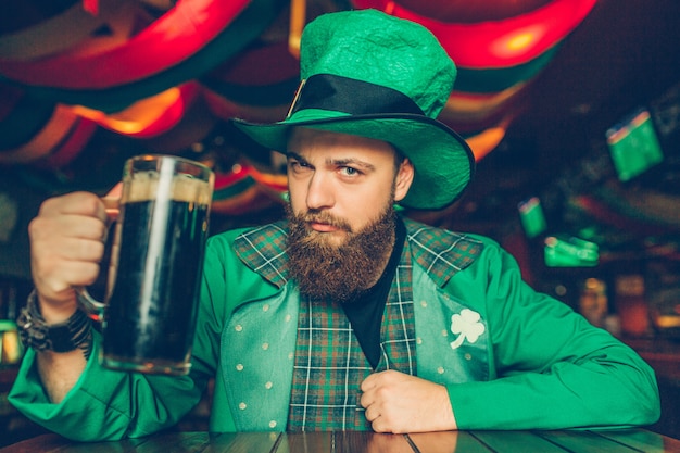 Seguro y serio joven en traje verde de San Patricio sentarse a la mesa en el pub y posar. Sostiene una jarra de cerveza oscura.