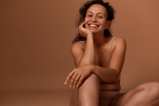 Seguro de sí mismo hermosa mujer de mediana edad en ropa interior beige, sonríe con dientes sonrisa mirando a la cámara. Concepto de positividad corporal, autoaceptación y amor corporal.