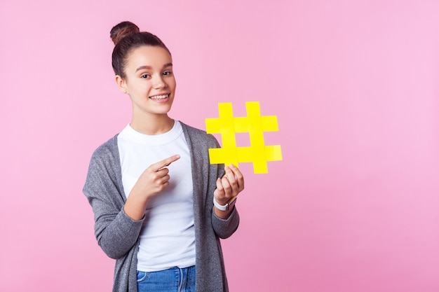 Sehen Sie sich beliebte Inhalte an Porträt eines trendigen brünetten Teenager-Mädchens mit Brötchenfrisur in Freizeitkleidung, das lächelt und auf ein großes gelbes Hashtag-Symbol zeigt Studioaufnahme isoliert auf rosa Hintergrund