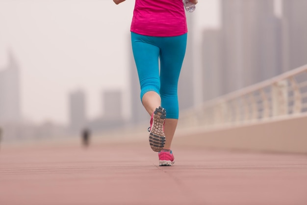 Sehr aktive junge schöne Frau, die damit beschäftigt ist, auf der Promenade am Meer entlang zu laufen, mit einer großen modernen Stadt im Hintergrund, um ihre Fitness so gut wie möglich aufrechtzuerhalten