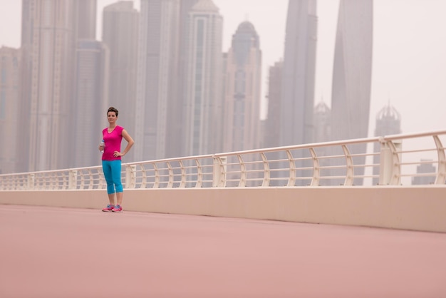 Sehr aktive junge schöne Frau, die damit beschäftigt ist, auf der Promenade am Meer entlang zu laufen, mit einer großen modernen Stadt im Hintergrund, um ihre Fitness so gut wie möglich aufrechtzuerhalten