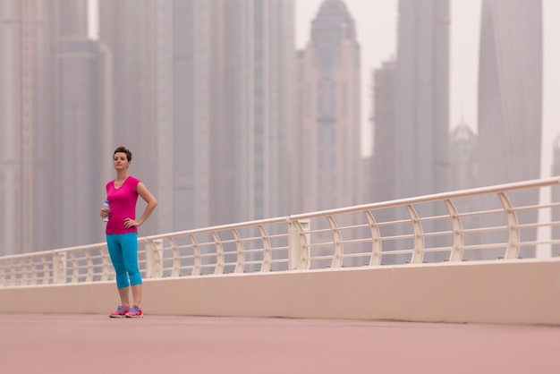 Sehr aktive junge schöne Frau, die damit beschäftigt ist, auf der Promenade am Meer entlang zu laufen, mit einer großen modernen Stadt im Hintergrund, um ihre Fitness so gut wie möglich aufrechtzuerhalten