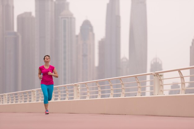 Sehr aktive junge schöne Frau, die damit beschäftigt ist, auf der Promenade am Meer entlang zu laufen, mit einer großen modernen Stadt im Hintergrund, um ihre Fitness so gut wie möglich aufrechtzuerhalten