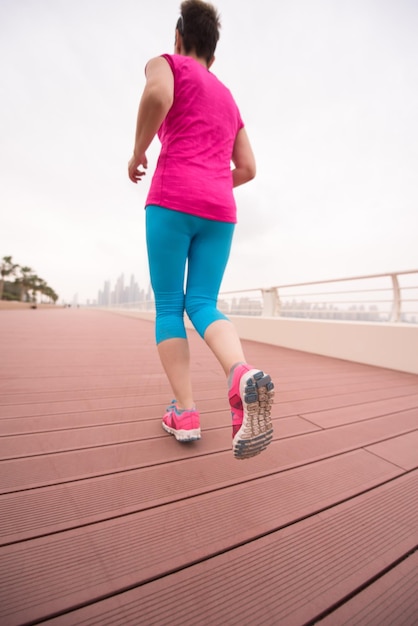 Sehr aktive junge schöne Frau, die damit beschäftigt ist, auf der Promenade am Meer entlang zu laufen, mit einer großen modernen Stadt im Hintergrund, um ihre Fitness so gut wie möglich aufrechtzuerhalten