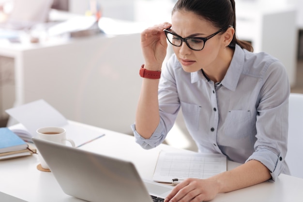 Sehr aufmerksame Arbeiterin, die ihre Stirn runzelt und die Brille berührt, während sie auf den Bildschirm ihres Laptops schaut