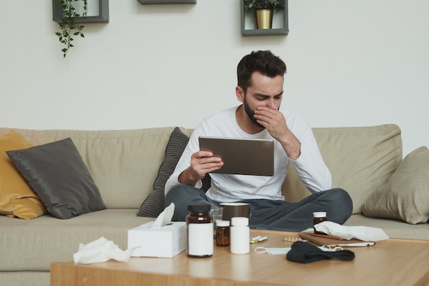 Sehr kranker junger Mann mit digitaler Tablette, die auf Couch sitzt und seinen Mund bedeckt, während er während der Fernarbeit in der häuslichen Umgebung hustet