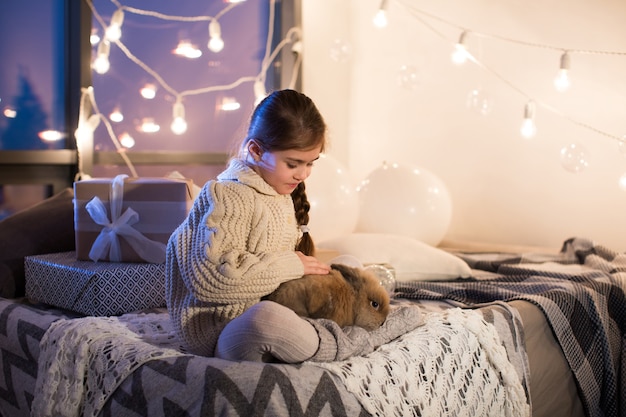 Sehr schönes und charmantes kleines dunkelhaariges Mädchen im weißen Pullover hält ein lebendes Kaninchen im Inneren des Hauses. Neujahr. Weihnachten. Hase. Studio.