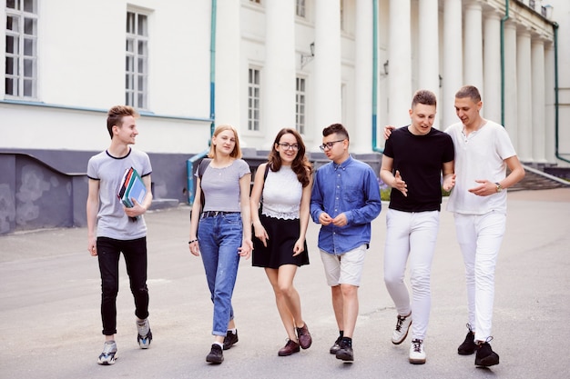 Seis alunos amigáveis caminham perto do prédio da universidade depois das aulas, sorrindo, aproveitando o tempo livre