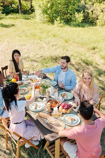 Seis jovens amigos de várias etnias reunidos à mesa servidos para o jantar, conversam e se deliciam com comida caseira em dia de verão