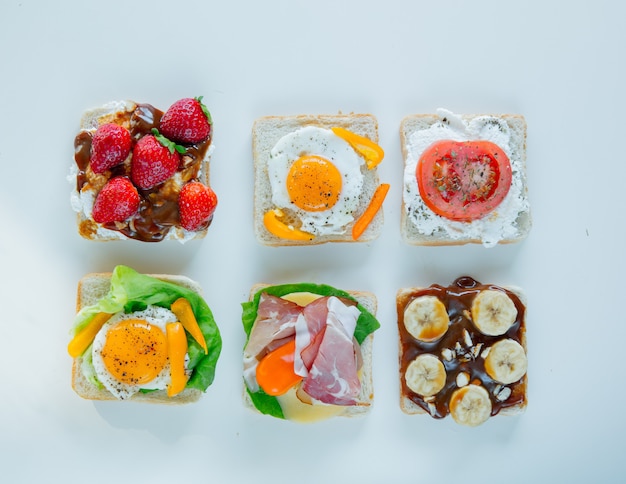 Seis sanduíches com tomate, omelete, chocolate, presunto e queijo na superfície branca. Acima vista